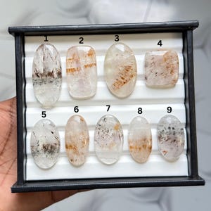 May include: A black display tray containing nine polished, translucent gemstones with internal inclusions. The stones are numbered one through nine, featuring various oval and rectangular shapes with clear, white, and reddish-brown mineral patterns.