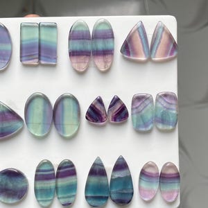 Pu&ograve; includere: Una collezione di pietre preziose di fluorite lucidata disposte in file su un espositore bianco. Le pietre presentano bande traslucide di colore viola, verde acqua e bianco trasparente in varie forme, tra cui ovali, rettangoli, triangoli e gocce.