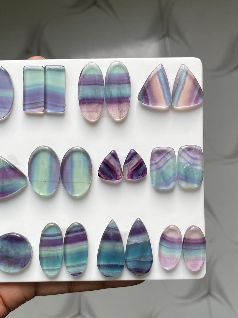 Pu&ograve; includere: Una collezione di pietre preziose di fluorite lucidata disposte in file su un espositore bianco. Le pietre presentano bande traslucide di colore viola, verde acqua e bianco trasparente in varie forme, tra cui ovali, rettangoli, triangoli e gocce.