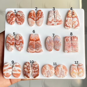May include: A selection of twelve pairs of polished gemstones, each with distinct patterns and colours. The stones display shades of red, orange, and cream, with intricate veining and swirls. The shapes vary, including ovals, triangles, and rectangles, presented on a white surface.