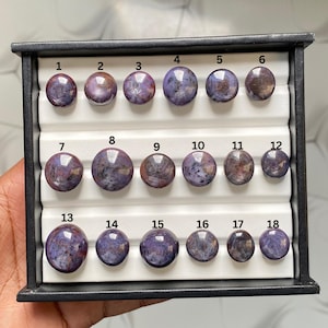 Peut inclure: Une boîte de présentation noire contenant dix-huit pierres précieuses cabochons rondes et polies dans des tons de violet, bleu et gris. Chaque pierre est numérotée de 1 à 18, disposée en trois rangées sur un insert blanc rainuré pour bijoux.