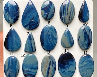 May include: A collection of fifteen polished cabochons arranged in a grid on a white surface. Each piece features a unique marbled pattern in shades of deep navy, royal blue, and light grey, with some stones displaying teardrop or oval shapes.