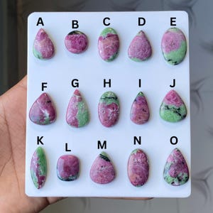 Könnte beinhalten: Eine Sammlung von fünfzehn polierten Rubin-Zoisit-Edelsteinen, jede mit einer einzigartigen Form und Muster. Die Steine zeigen eine Mischung aus rosa, grünen und schwarzen Farben. Die Edelsteine sind auf einer weißen Oberfläche angeordnet und mit Buchstaben beschriftet.