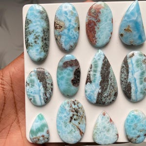 May include: A collection of eleven polished Larimar cabochons in various shapes including ovals and teardrops. Each stone features a unique pattern of sky blue, white, and dark brown or black mineral inclusions, displayed on a white board.