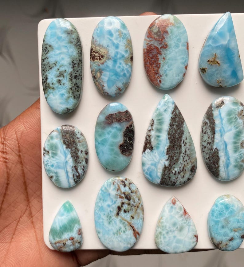 May include: A collection of eleven polished Larimar cabochons in various shapes including ovals and teardrops. Each stone features a unique pattern of sky blue, white, and dark brown or black mineral inclusions, displayed on a white board.