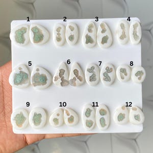 May include: A collection of twelve pairs of white cabochons arranged on a white display card, numbered one through twelve. Each cabochon features a smooth, opaque white border surrounding a central, textured, light green and tan mineral inclusion.