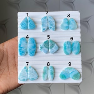 May include: Nine pairs of polished larimar gemstones in various shapes and sizes, displayed on a white grid. The gemstones range in color from light to medium blue, with white marbling patterns. The shapes include teardrops, ovals, and irregular forms.