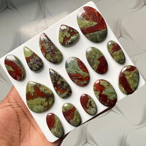 May include: A collection of fifteen polished cabochons featuring a mix of deep red and olive green patterns. These smooth, rounded stones are arranged on a white display board, showcasing varied shapes including ovals and teardrops with glossy surfaces.