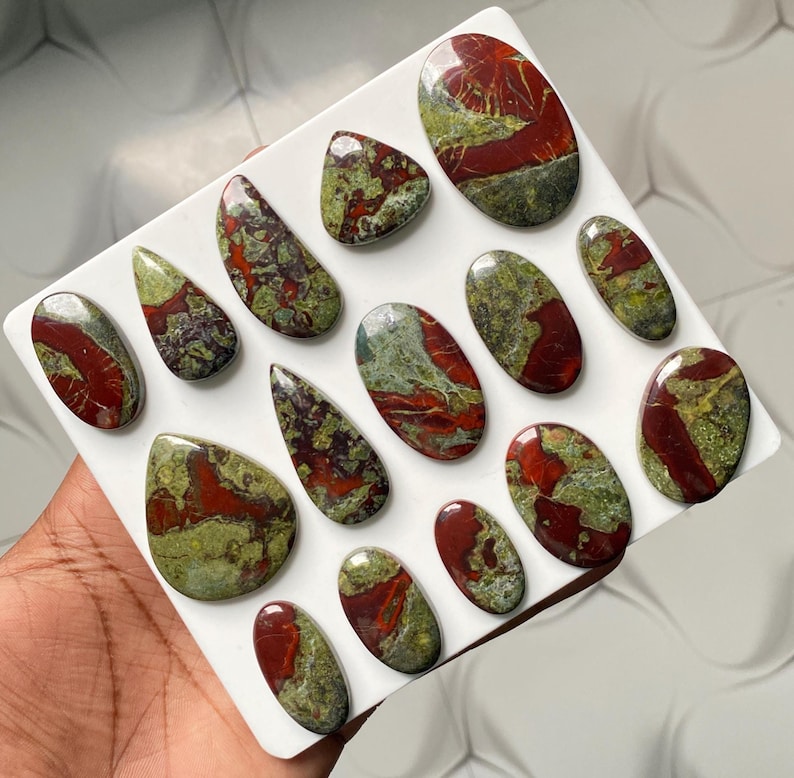 May include: A collection of fifteen polished cabochons featuring a mix of deep red and olive green patterns. These smooth, rounded stones are arranged on a white display board, showcasing varied shapes including ovals and teardrops with glossy surfaces.