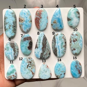 May include: A collection of fifteen polished larimar cabochons arranged on a white display board. Each stone features unique patterns of sky blue, white, and brown inclusions in various shapes including ovals, teardrops, and rounded forms, numbered 1-15.