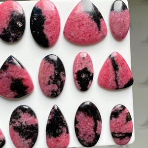May include: A collection of polished rhodonite cabochons in various shapes including ovals, triangles, and teardrops. Each stone features a distinct pattern of vibrant pink and deep black mineral inclusions with a smooth, glossy surface finish.