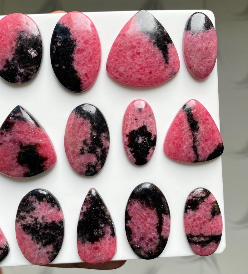 May include: A collection of polished rhodonite cabochons in various shapes including ovals, triangles, and teardrops. Each stone features a distinct pattern of vibrant pink and deep black mineral inclusions with a smooth, glossy surface finish.