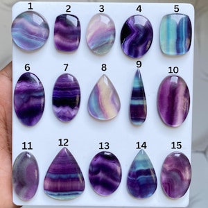 May include: Fifteen polished fluorite gemstones in various shapes, including oval, teardrop, and rectangular. The stones display a mix of purple, blue, and white colours. The gemstones are arranged on a white surface.