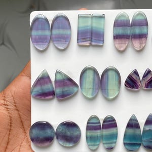 Pu&ograve; includere: Una collezione di cabochon in fluorite lucida in varie forme, tra cui ovali, rettangoli, triangoli, cerchi e gocce. Ogni pietra presenta distinte bande orizzontali di colore viola, verde acqua e verde, disposte in coppia su un supporto bianco.