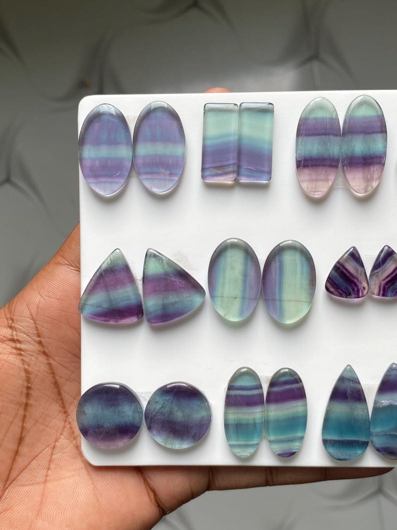 Pu&ograve; includere: Una collezione di cabochon in fluorite lucida in varie forme, tra cui ovali, rettangoli, triangoli, cerchi e gocce. Ogni pietra presenta distinte bande orizzontali di colore viola, verde acqua e verde, disposte in coppia su un supporto bianco.