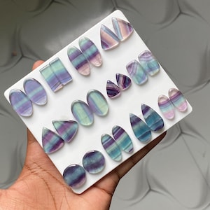 Pu&ograve; includere: Una collezione di cabochon in fluorite lucida di varie forme, tra cui ovali, triangoli e gocce, disposti su un supporto bianco. Ogni pietra presenta bande distinte di strati minerali di colore viola, blu, verde e bianco traslucido.