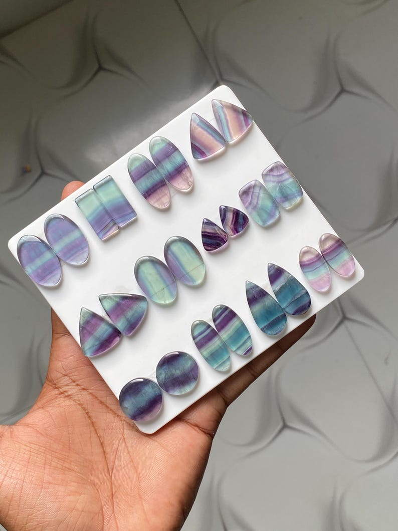 Pu&ograve; includere: Una collezione di cabochon in fluorite lucida di varie forme, tra cui ovali, triangoli e gocce, disposti su un supporto bianco. Ogni pietra presenta bande distinte di strati minerali di colore viola, blu, verde e bianco traslucido.