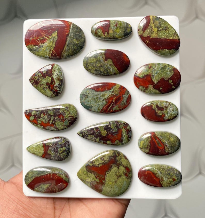 May include: A collection of fifteen polished cabochons arranged on a white rectangular display board. Each stone features a distinct pattern of deep red and olive green mineral inclusions with a smooth, glossy finish in various oval and teardrop shapes.