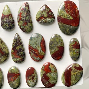 May include: A collection of polished cabochons featuring a mix of deep red and olive green patterns. These stones are shaped into various ovals, teardrops, and rounded forms, each displaying a smooth, glossy surface with unique mineral inclusions.
