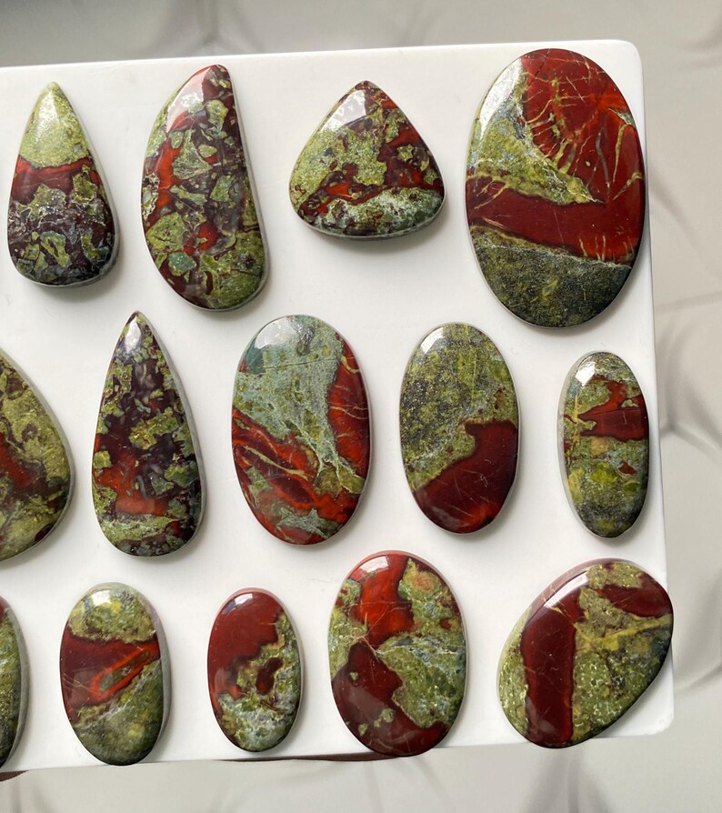 May include: A collection of polished cabochons featuring a mix of deep red and olive green patterns. These stones are shaped into various ovals, teardrops, and rounded forms, each displaying a smooth, glossy surface with unique mineral inclusions.