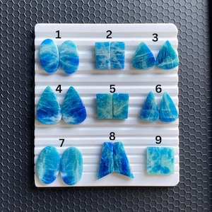 May include: A collection of nine pairs of polished blue gemstones in various shapes, including ovals, squares, teardrops, and triangles. The gemstones are displayed on a white tray with a ridged surface, set against a dark background.