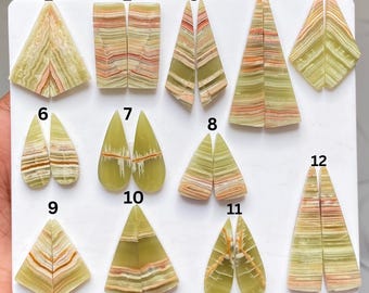 Può includere: Una collezione di dodici paia di cabochon in onice verde lucido, numerati da uno a dodici. Queste pietre presentano motivi a bande orizzontali e angolari in tonalità verde chiaro, crema e arancione, disposte su una superficie bianca.
