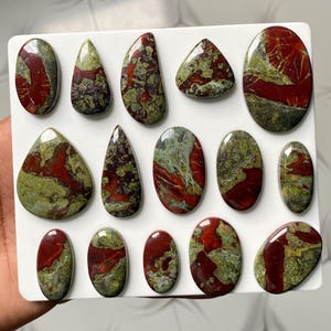 May include: A collection of fourteen polished cabochons featuring a mottled pattern of deep red and olive green. These stones are arranged in rows on a white display board, showcasing various shapes including ovals, teardrops, and rounded forms.