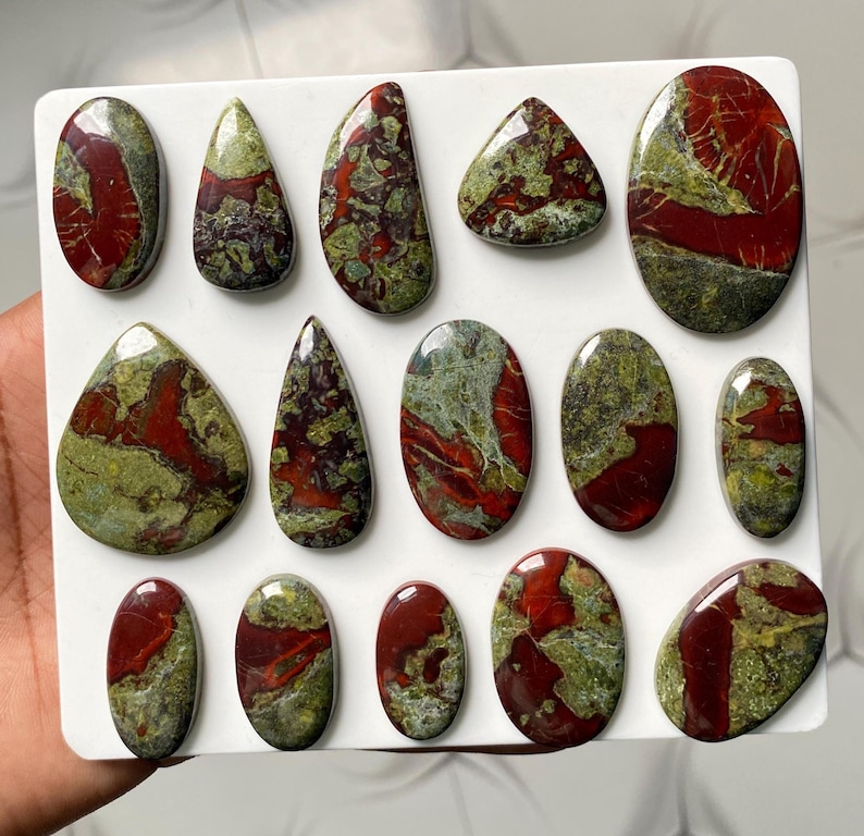 May include: A collection of fourteen polished cabochons featuring a mottled pattern of deep red and olive green. These stones are arranged in rows on a white display board, showcasing various shapes including ovals, teardrops, and rounded forms.