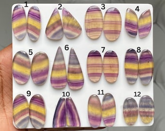 May include: A collection of twelve pairs of polished, multi-colored fluorite cabochons arranged on a white display board. Each stone features horizontal bands of purple, yellow, and translucent white. The stones are numbered 1 through 12.