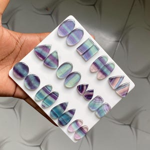 Pu&ograve; includere: Una collezione di cabochon in fluorite lucida di varie forme, tra cui ovali, triangoli e gocce, esposti su un supporto quadrato bianco. Le pietre presentano motivi a bande in tonalit&agrave; viola, verde, blu e bianco traslucido.