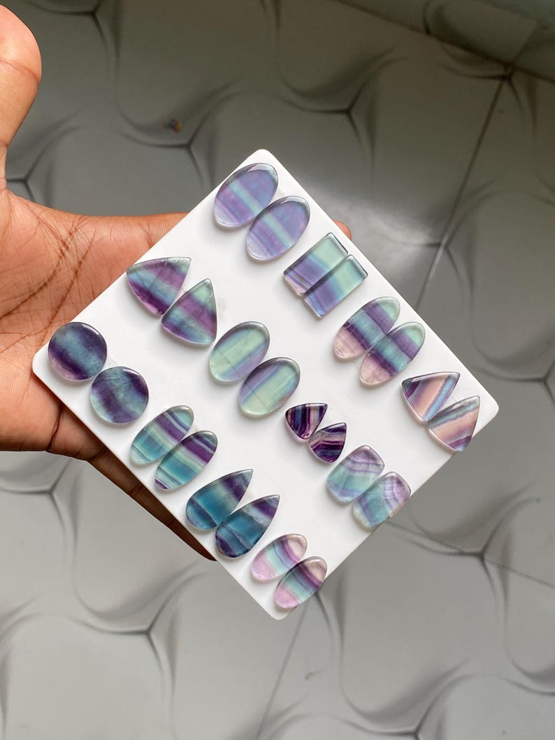 Pu&ograve; includere: Una collezione di cabochon in fluorite lucida di varie forme, tra cui ovali, triangoli e gocce, esposti su un supporto quadrato bianco. Le pietre presentano motivi a bande in tonalit&agrave; viola, verde, blu e bianco traslucido.