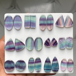 Pu&ograve; includere: Una collezione di cabochon in fluorite lucida disposti su un espositore bianco. Le pietre presentano motivi a bande nei toni del viola, blu, verde e bianco traslucido, sagomate in ovali, rettangoli, triangoli, cerchi e forme a goccia.