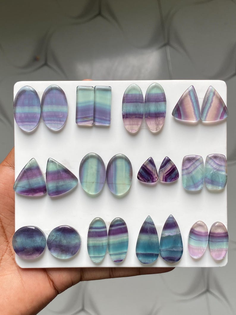 Pu&ograve; includere: Una collezione di cabochon in fluorite lucida disposti su un espositore bianco. Le pietre presentano motivi a bande nei toni del viola, blu, verde e bianco traslucido, sagomate in ovali, rettangoli, triangoli, cerchi e forme a goccia.