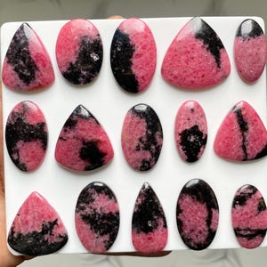 May include: A collection of fourteen polished rhodonite cabochons arranged on a white square display board. Each stone features a unique pattern of vibrant pink and deep black mineral inclusions, shaped into various ovals, triangles, and teardrops.