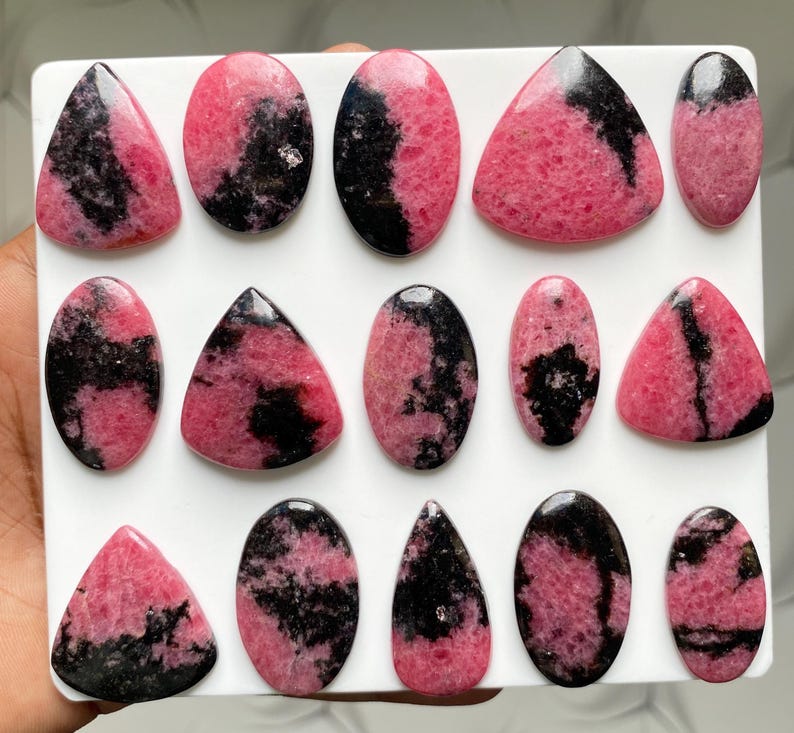 May include: A collection of fourteen polished rhodonite cabochons arranged on a white square display board. Each stone features a unique pattern of vibrant pink and deep black mineral inclusions, shaped into various ovals, triangles, and teardrops.