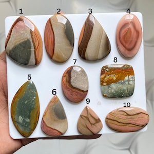 May include: Ten polished, multi-coloured jasper cabochons arranged on a white square board, numbered one through ten. Each stone features unique natural banding, swirls, and earthy tones including shades of pink, brown, grey, green, and tan.