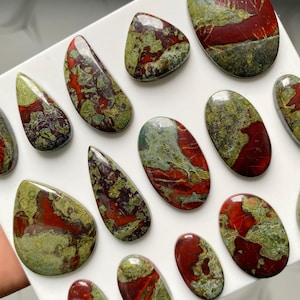 May include: A collection of polished, smooth cabochon gemstones featuring a marbled pattern of deep red and olive green. These stones are shaped into various ovals, teardrops, and rounded forms, all displaying a glossy, reflective surface finish.