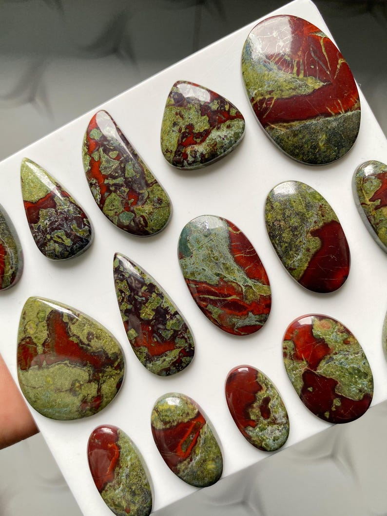 May include: A collection of polished, smooth cabochon gemstones featuring a marbled pattern of deep red and olive green. These stones are shaped into various ovals, teardrops, and rounded forms, all displaying a glossy, reflective surface finish.