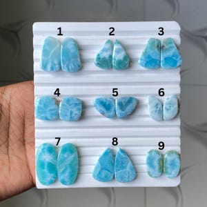 May include: Nine pairs of polished larimar gemstones, each displaying unique blue and white patterns. The stones are presented on a white, ridged surface, highlighting their diverse shapes and sizes, ideal for jewellery making.