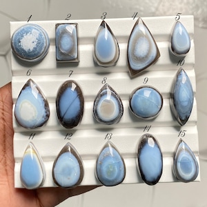 May include: A collection of fifteen polished blue and white cabochon gemstones arranged in three rows on a white display board. Each stone is numbered from one to fifteen and features unique banded patterns, smooth surfaces, and varied shapes.