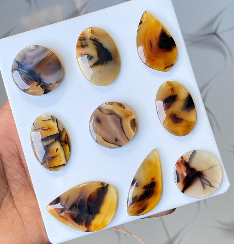 May include: A collection of polished agate gemstones in various shapes, including oval, teardrop, and round. The stones display a mix of warm colors, with shades of brown, tan, and yellow, and intricate patterns. The gemstones are arranged on a white surface.