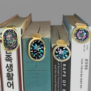 May include: Gold-toned metal bookmarks with inlaid iridescent floral designs are displayed on the spines of several books. The bookmarks have a decorative top and an oval or circular design. The books have text in English and Korean.