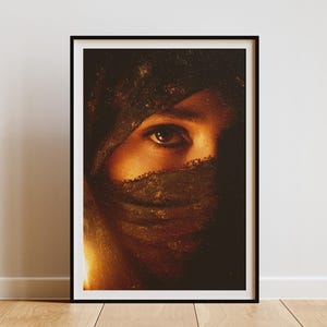 Puede incluir: Impresión artística enmarcada que presenta un primer plano de un ojo parcialmente velado. La obra de arte utiliza tonos cálidos y terrosos, centrándose en el ojo para crear una sensación de misterio. La impresión está en un marco negro, exhibida contra una pared blanca.