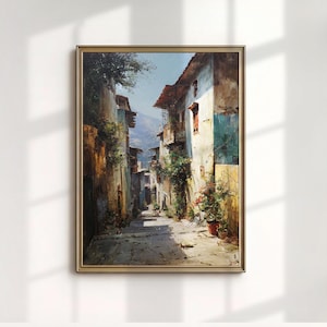 May include: A framed oil painting of a European street scene. The buildings are aged with textured walls in shades of brown, beige, and blue. Balconies and potted plants add detail. The sky is a clear blue, and the painting is in a gold frame.