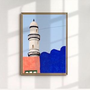 May include: Framed art print featuring a white minaret with black detailing against a light blue sky. The minaret is set against a coral-colored wall and a deep blue structure. A green window is visible at the base.