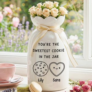 May include: A white ceramic jar with a scalloped rim, tied with a cream ribbon, containing light pink roses. The jar features the black text "YOU'RE THE SWEETEST COOKIE IN THE JAR" above illustrations of a cookie and a heart with faces, and the names "Lily" and "Sara".