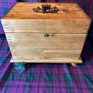 May include: A wooden chest with a carved emblem on the lid and a small keyhole. The chest is light brown and sits on a base with small, decorative feet. The background features a purple and green plaid fabric.