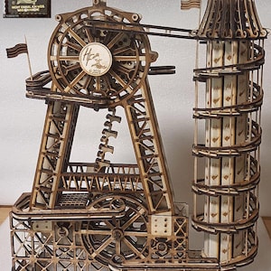Può includere: Un modello meccanico di pista per biglie in legno tagliato al laser con una grande ruota circolare, una torre a spirale e complesse strutture a ingranaggi. Il design include una ruota centrale con il testo 'Hf 2026' e un cartello 'HeFra Tower'.