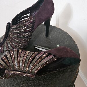 Może przedstawiać: Para fioletowych sandałów na wysokim obcasie z paskami. Sandały mają materiał przypominający zamsz na pięcie i palcach, ze srebrnymi i fioletowymi zdobieniami. Obcasy są czarne, a buty są prezentowane na ciemnoszarej powierzchni.