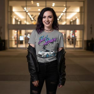 Peut inclure: Un t-shirt gris avec le texte "Just Drive" en turquoise et violet, et un dessin d'une voiture de sport blanche avec des accents bleus. Le t-shirt est porté par une personne portant une veste en cuir noire et un jean noir.